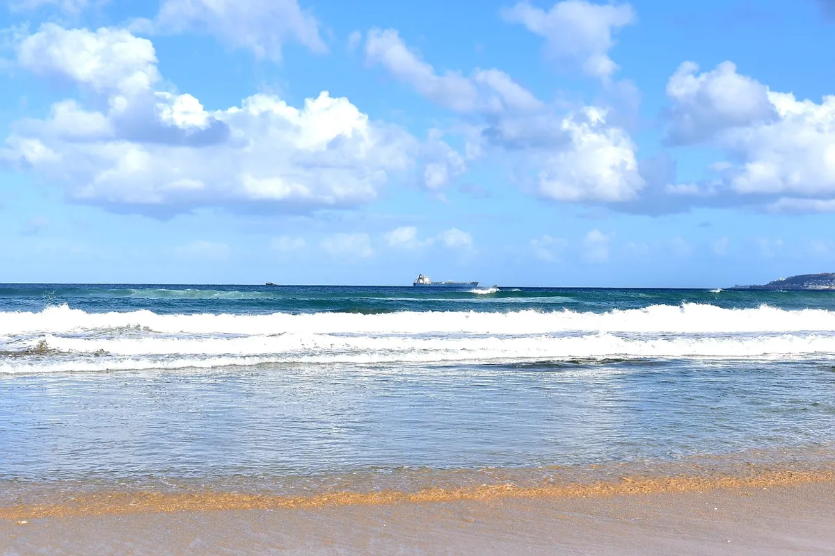 Successive lines of white surf breaking on the shore at Diaz Beach, Mossel Bay, with a cargo ship on the horizon under a blue sky