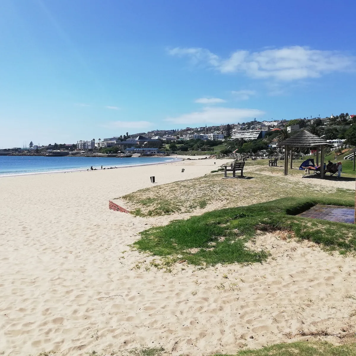 Santos Beach Mossel Bay — wide pale sand meeting flat turquoise water, thatched shade shelters and a bench on the grassed terrace above the beach, town buildings on the hillside beyond