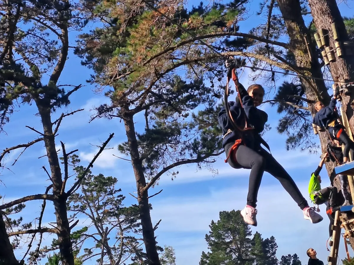 A girl in a harness riding a short zipline between pine trees, with other climbers on a platform behind