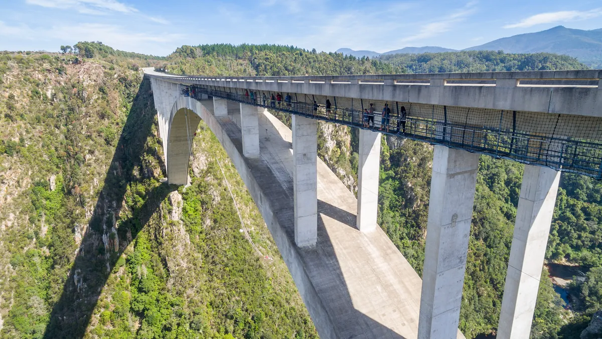 The concrete arch of the Bloukrans Bridge spanning a forested gorge, with people standing on the walkway under the deck