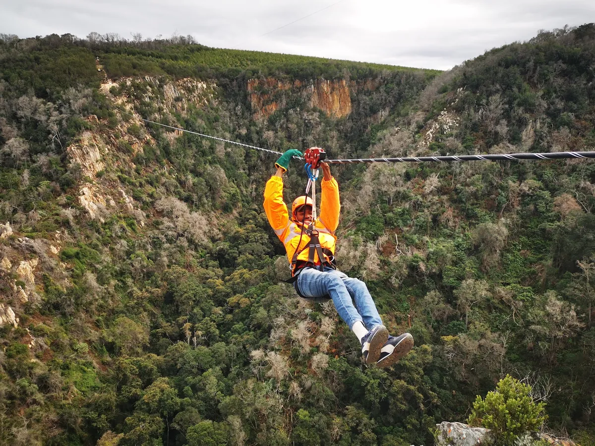 A smiling rider in an orange jacket and helmet on a zipline cable above the forested Kranshoek gorge