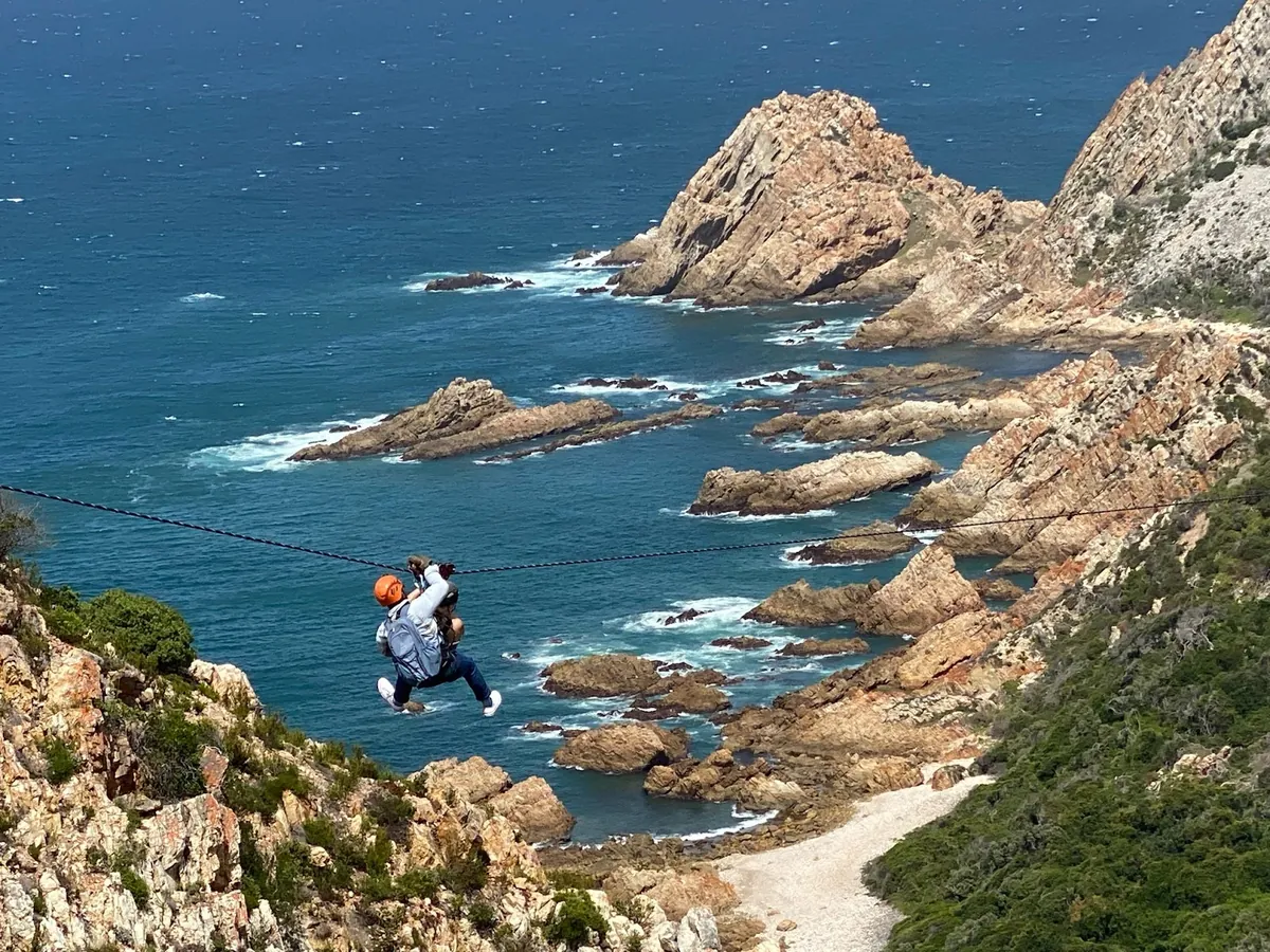 A rider in an orange helmet on a zipline above rocky sea stacks and blue water at Kranshoek, near Plettenberg Bay