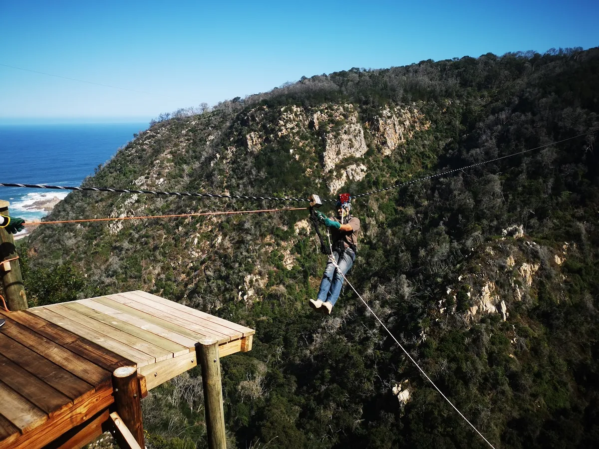 A rider leaving a wooden platform on a cable across a gorge, with the sea on the left