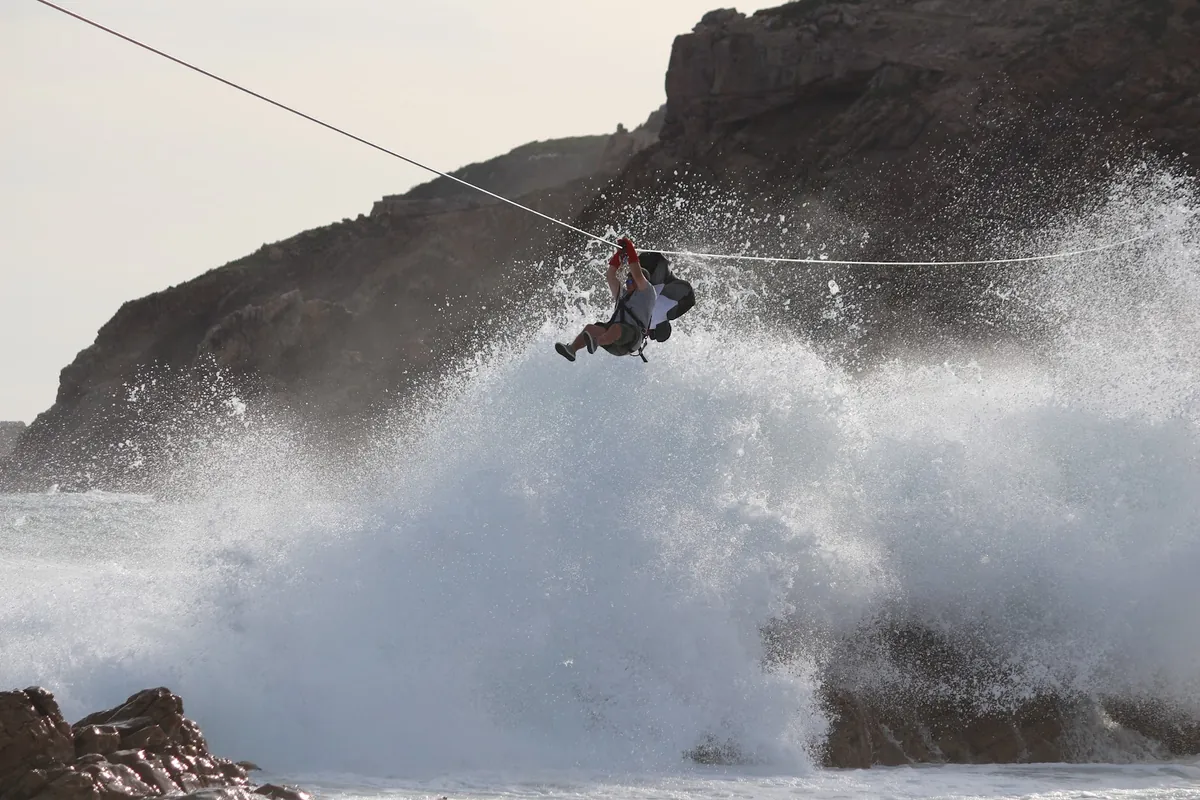 A rider on the Mossel Bay Zipline cable as a wave breaks into spray on the rocks below the cliffs