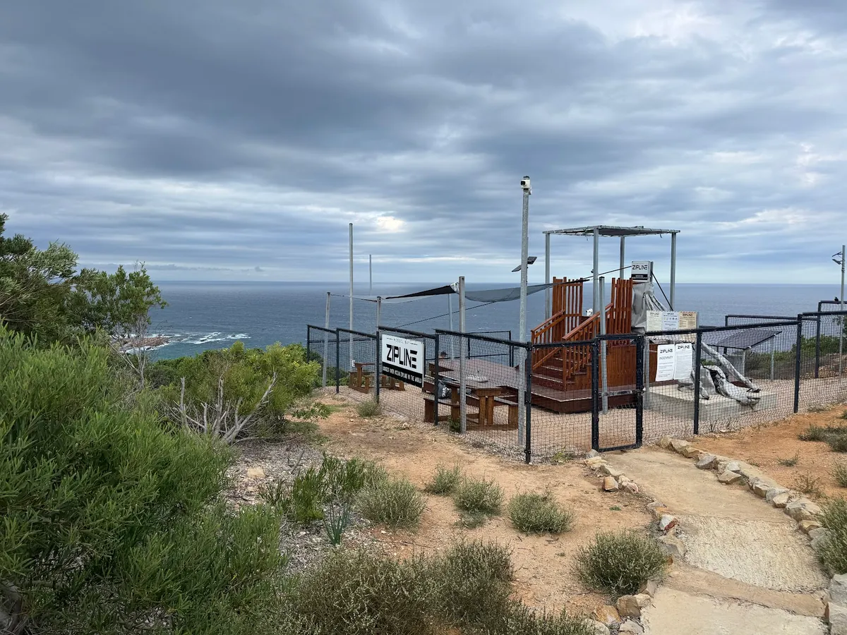 The fenced Mossel Bay Zipline start platform on a fynbos-covered clifftop, with the ocean behind under a grey sky
