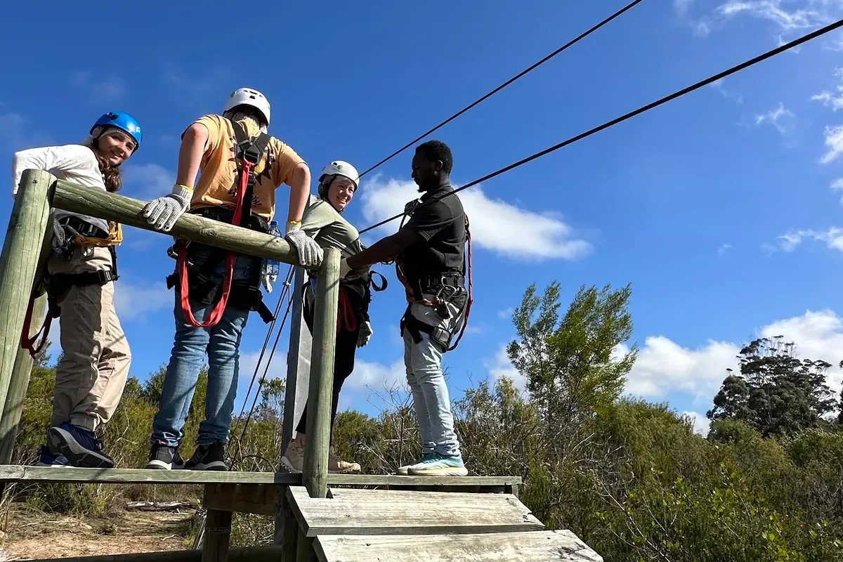 Four people in helmets and harnesses on a wooden zipline platform with two cables running off into a blue sky
