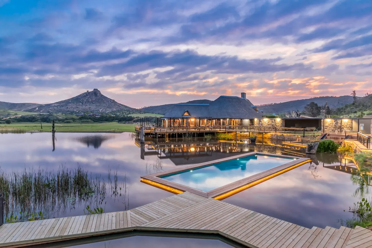 Botlierskop's thatched main lodge and infinity pool reflected in the reserve dam at dusk, with a rocky peak behind
