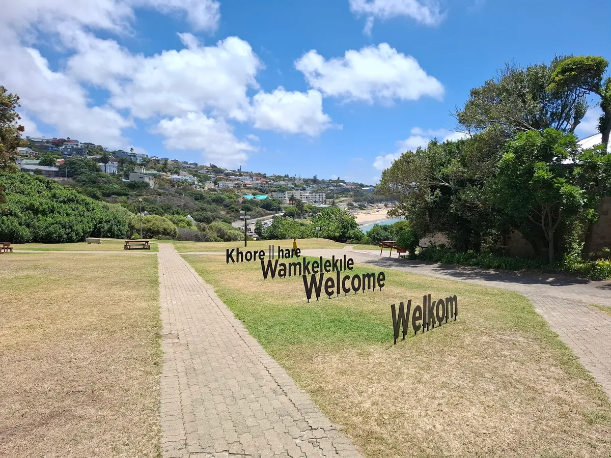 The multilingual Welcome, Welkom, Wamkelekile signs on a grassy rise above Mossel Bay with the Indian Ocean and Santos Bay in the background