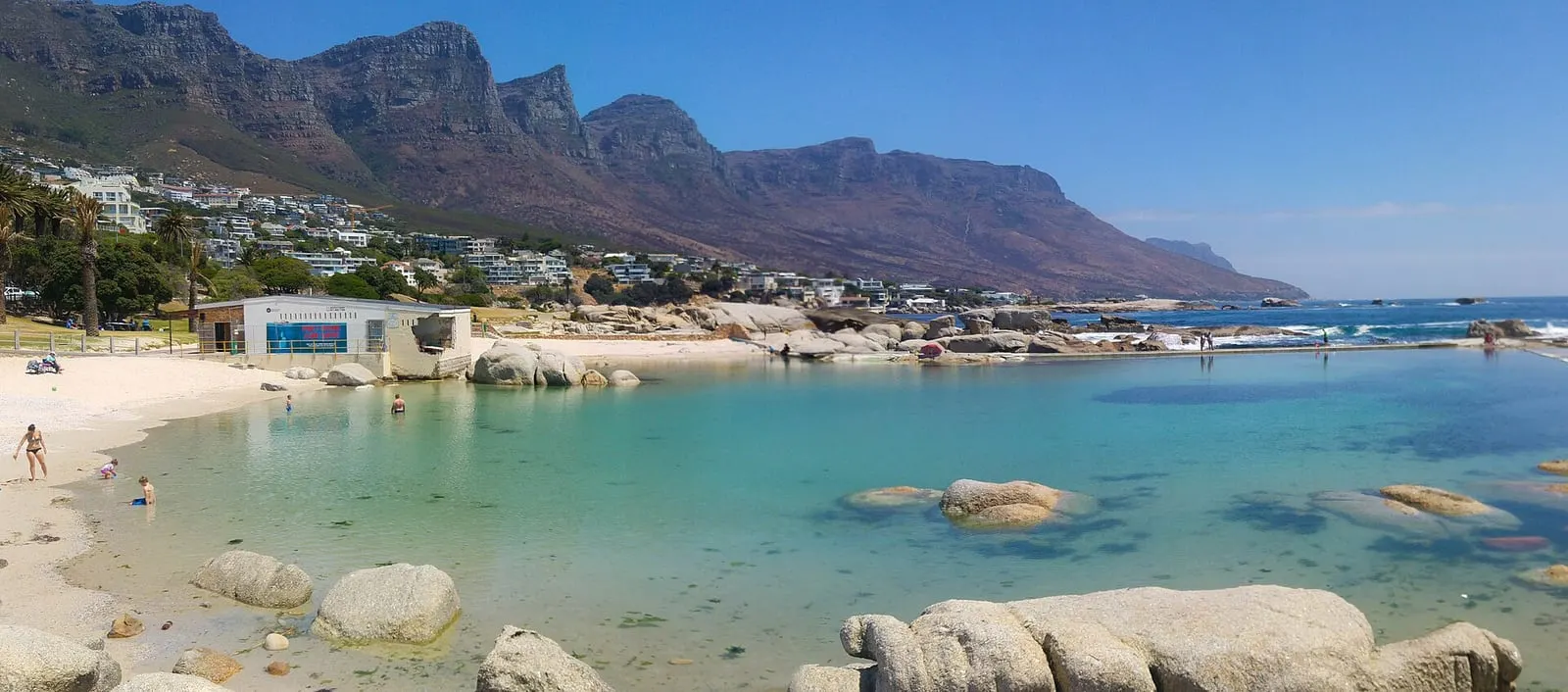 The Camps Bay tidal pool on a clear day: shallow turquoise water between granite boulders, a few bathers, the Twelve Apostles behind