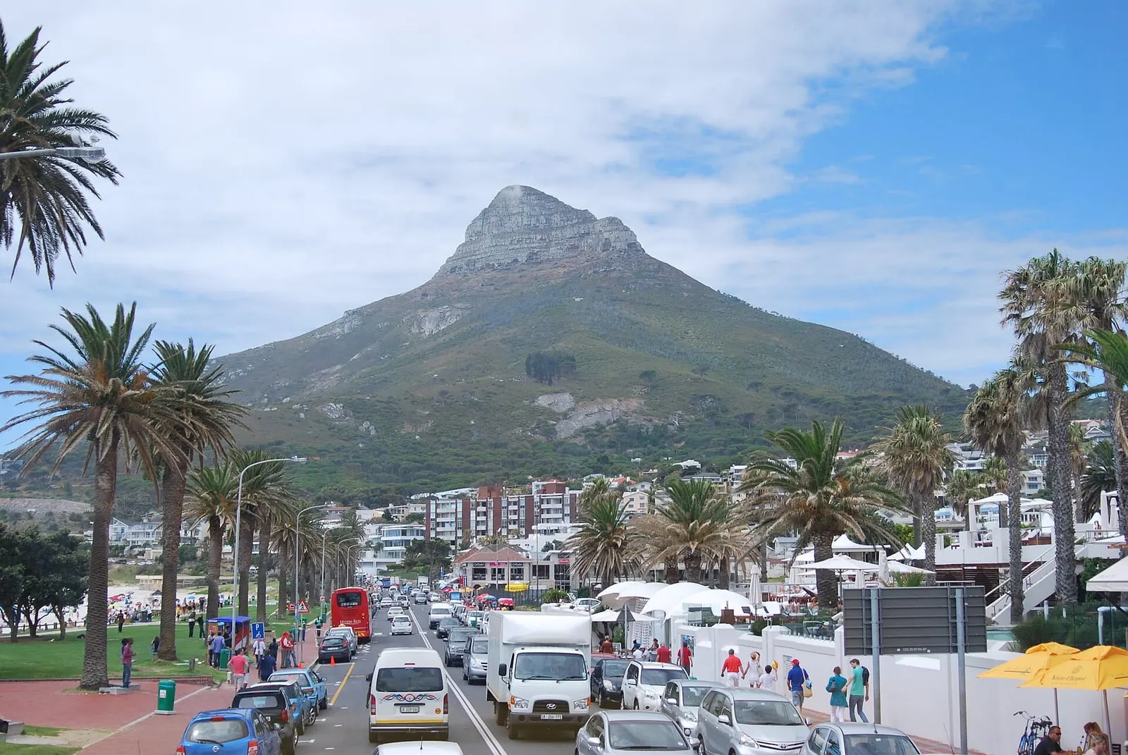 Victoria Road in Camps Bay lined with palms and traffic, beachgoers on the lawn and Lion's Head filling the sky